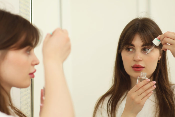 Woman applying a morning skincare serum in front of a mirror in warm natural light